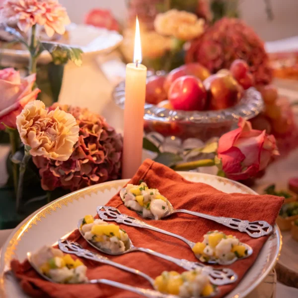 beautiful antique spoons filled iwth ceviche displayed on a napkin on a plate, candle and flowers of the same colors are making this shot a classical beauty catering offer.